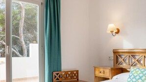 A bedroom with a bed, a nightstand, a chair, a wall-mounted lamp, and a sliding glass door leading to an outdoor area. - Rocaplana Hostal (Formentera)