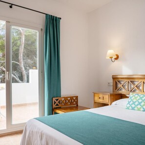 A bedroom with a bed, a nightstand, a chair, a wall-mounted lamp, and a sliding glass door leading to an outdoor area. - Rocaplana Hostal (Formentera)