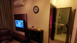 Une chambre d’hôtel équipée d’une télévision, d’une horloge, d’une cabine de douche en verre et d’un meuble en bois foncé.