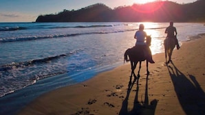 Una spiaggia con due persone a cavallo, un tramonto sullo sfondo e onde che si infrangono sulla riva.
