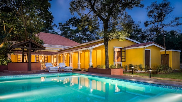 A pool area with a clear blue pool, lounge chairs, and a yellow building with a red roof. - Ama Stays And Trails Cardozo House (Candolim)