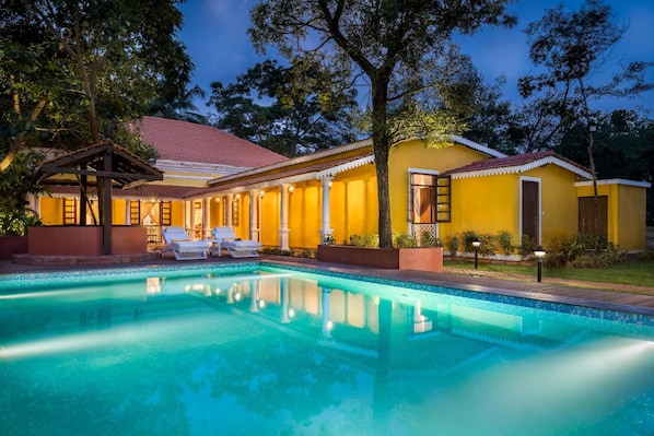 A pool area with a clear blue pool, lounge chairs, and a yellow building with a red roof. - Ama Stays And Trails Cardozo House (Candolim)