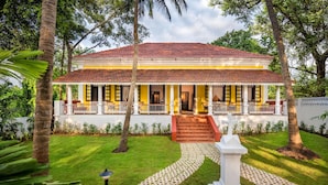 A two-story building with a red-tiled roof, a porch with white railings, and a garden with a white statue. - Ama Stays And Trails Cardozo House (Candolim)