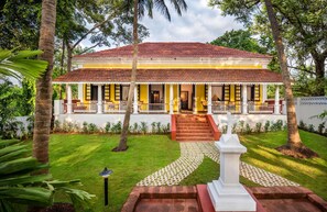 A two-story building with a red-tiled roof, a porch with white railings, and a garden with a white statue. - Ama Stays And Trails Cardozo House (Candolim)