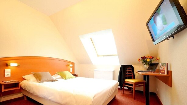 A hotel room with a bed, a desk with a laptop, a television mounted on the wall, and a window providing natural light. - Comfort Hotel Emerainville Marne-la-vallee (Emerainville)