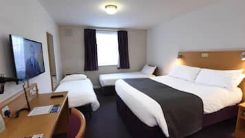 A hotel room with a bed, two smaller beds, a desk with a chair, a television, and a window with curtains.
