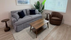 A modern living room with a grey sofa, a wooden coffee table, a brown armchair, a ceiling fan, and a window with blinds. - 18 On the Bay Suite #6 (Indian Rocks Beach)