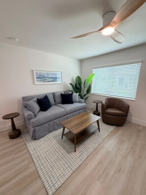 A modern living room with a grey sofa, a wooden coffee table, a brown armchair, a ceiling fan, and a window with blinds. - 18 On the Bay Suite #6 (Indian Rocks Beach)