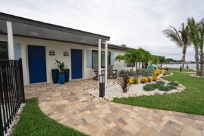 Een modern gebouw met een verharde opgang, een blauwe deur en een aangelegde tuin met palmbomen en gele bloemen.