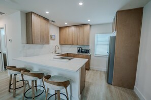 Een moderne keuken met houten kastjes, een wit aanrecht, een ingebouwde magnetron en een koelkast. Drie barkrukken met beklede zittingen.