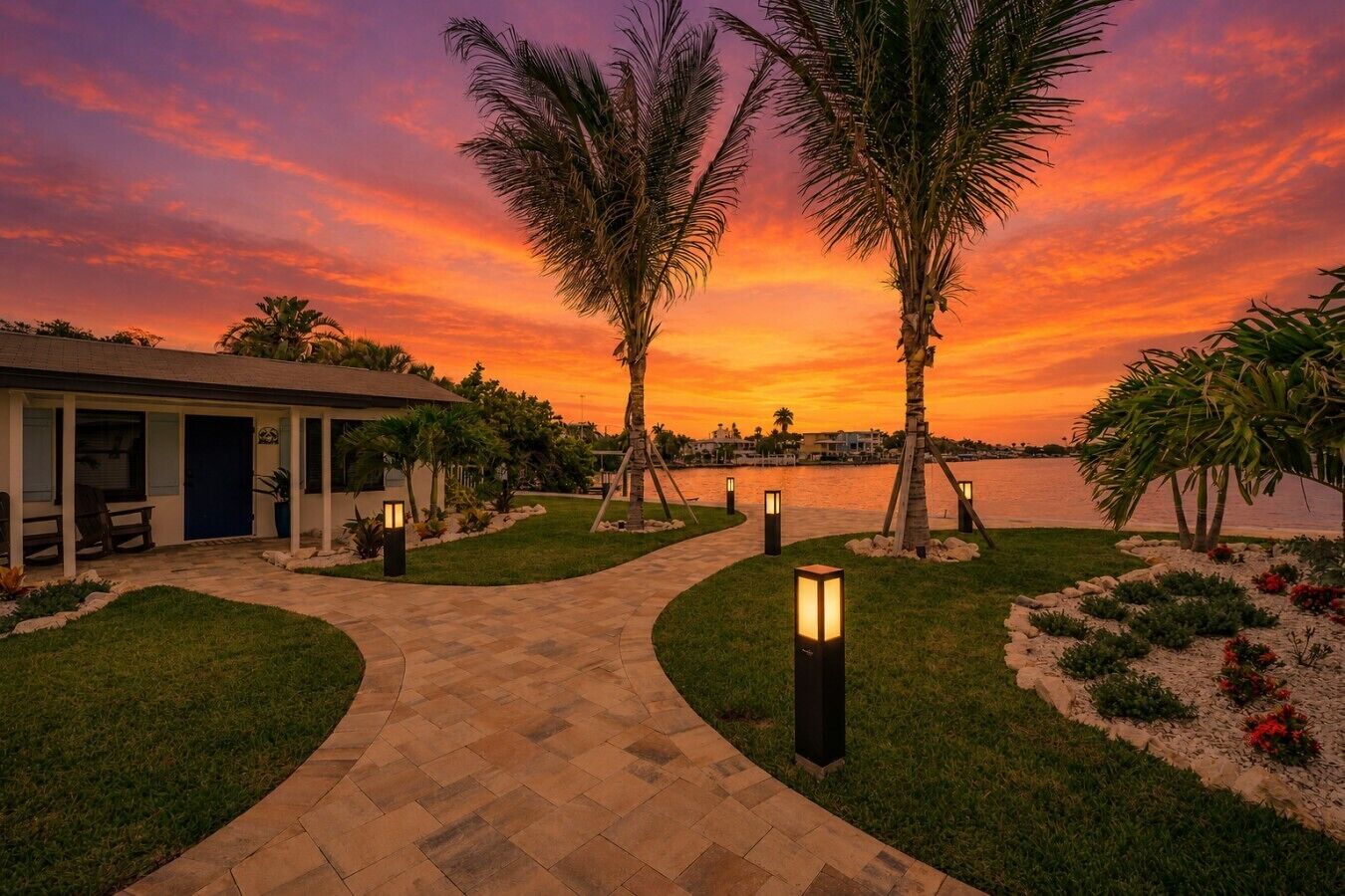 Un camino que lleva a un edificio con vistas a un atardecer sobre el agua, palmeras e iluminación exterior.