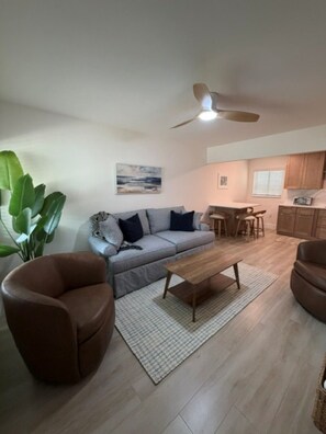 Een woonkamer met een bank, salontafel en twee fauteuils, een eethoek met tafel en stoelen, en een keuken met kasten en een raam.