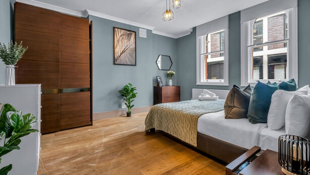 Un dormitorio moderno con un armario de madera, una cama con edredón blanco, una mesita de noche con lámpara y una ventana con vistas al exterior.