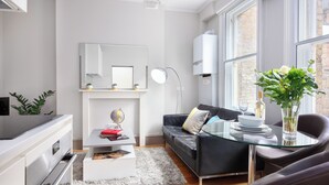 Una sala moderna con una mesa de centro de vidrio, un sofá negro, una chimenea blanca y un ventanal con vistas a un edificio de ladrillo.