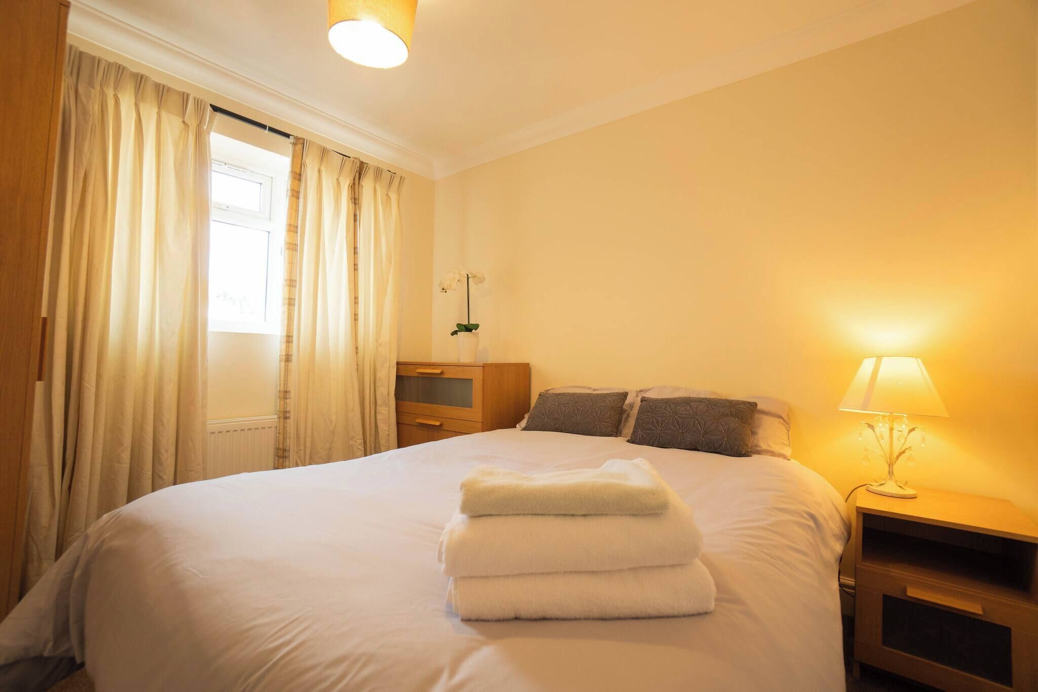 Ein Hotelzimmer mit Bett, Nachttisch, Lampe, einem Fenster mit Vorhängen und einer Heizung.