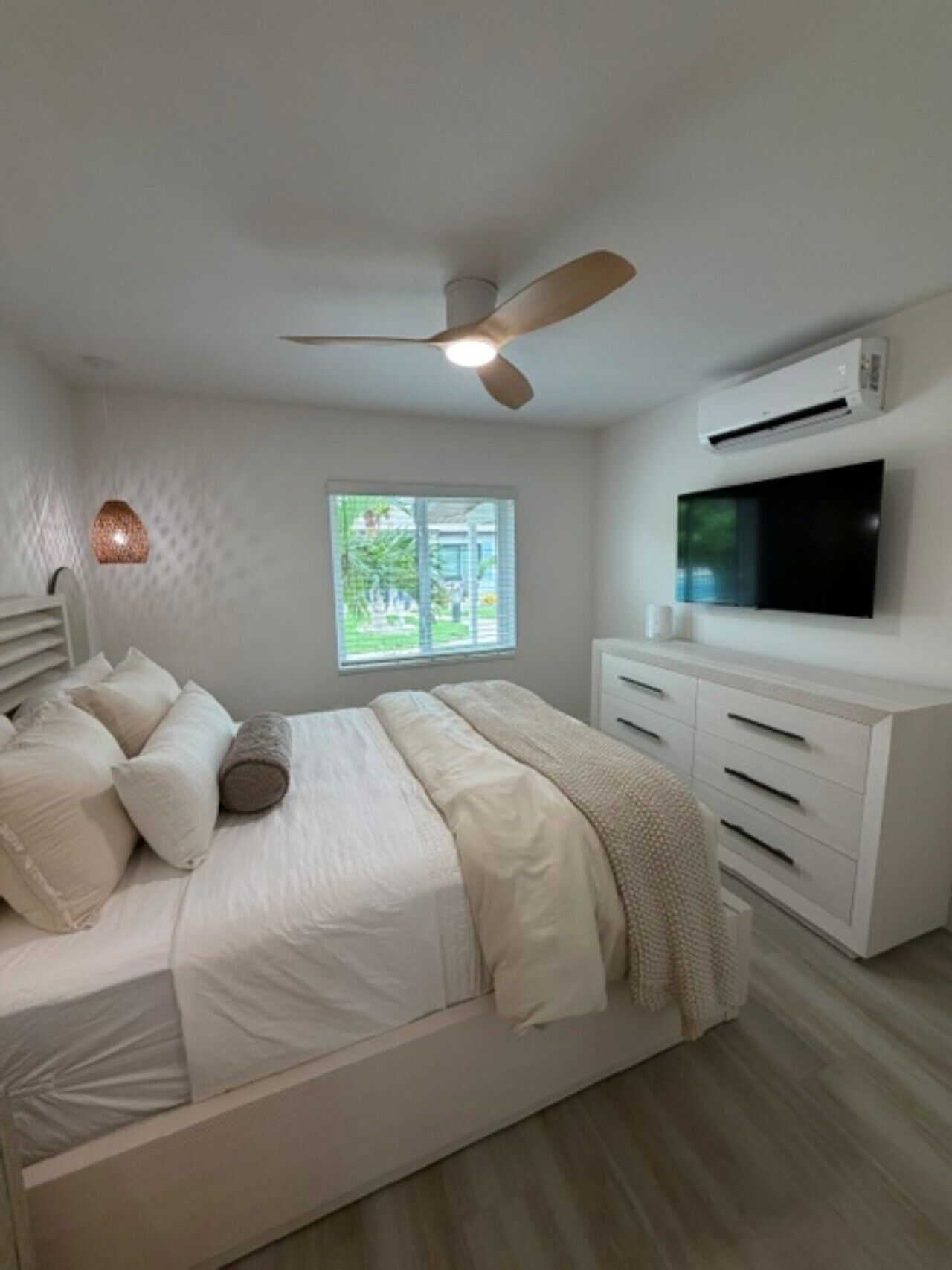 A bedroom with a bed, a ceiling fan, an air conditioning unit, a TV, and a window with a view of greenery.