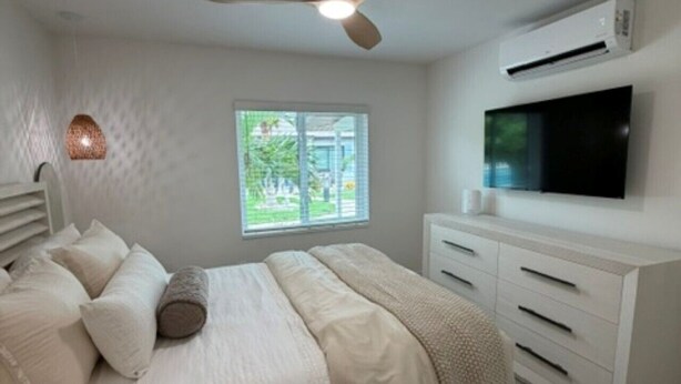 Un dormitorio con cama, ventilador de techo, aire acondicionado, televisor y ventana con vista a follaje.