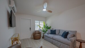 Una sala de estar con sofá, dos sillones, ventilador de techo, televisor y ventana con planta.