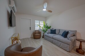 Ein Wohnzimmer mit einer Couch, zwei Sesseln, einem Deckenventilator, einem Fernseher und einem Fenster mit einer Pflanze.