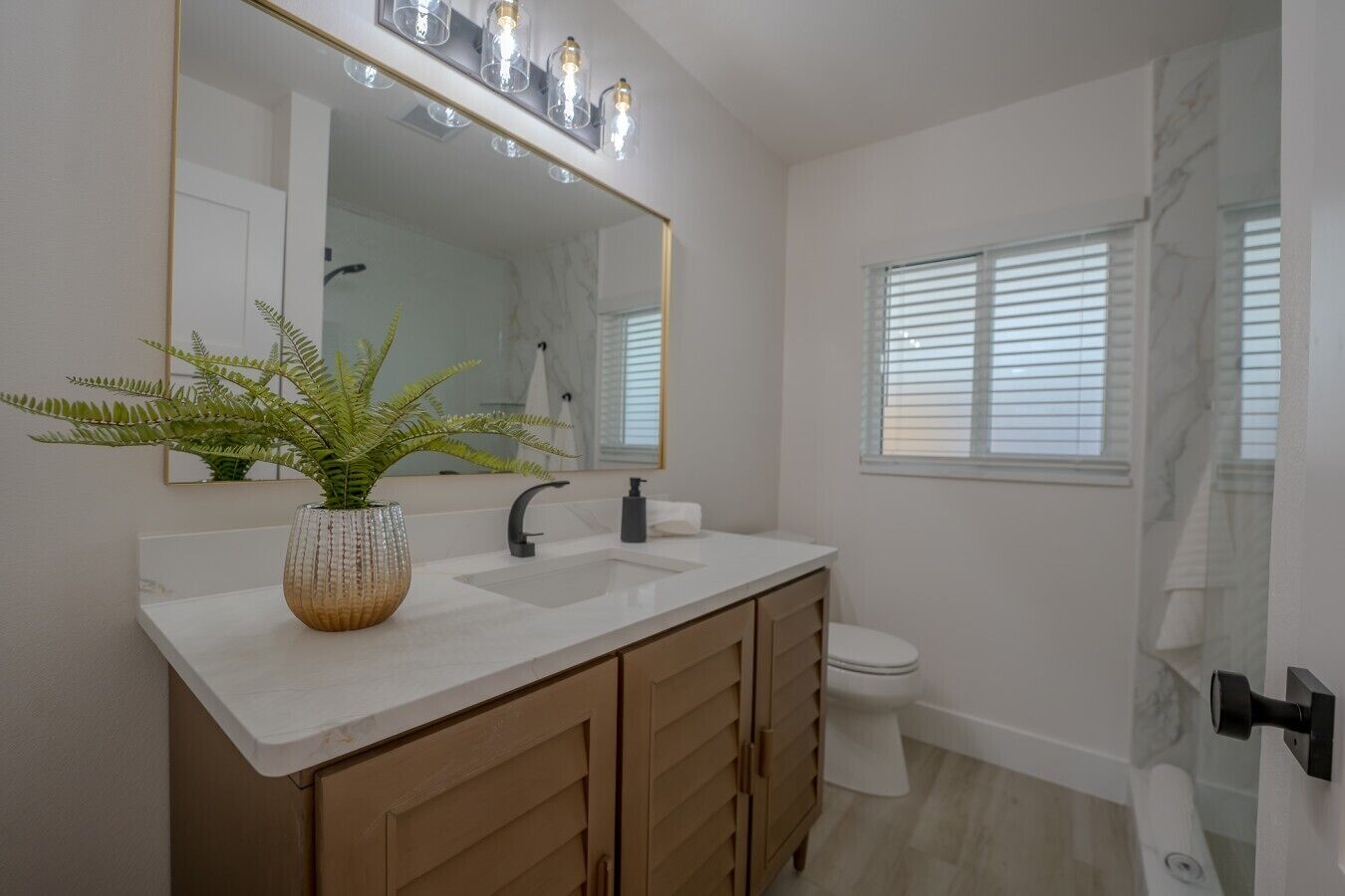 A bathroom with a white sink, a mirror with lights, a toilet, and a window with blinds.