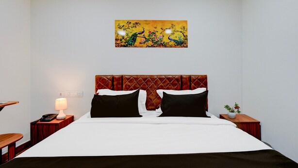 A hotel room with a bed, a nightstand with a lamp, a wall-mounted painting, and a small table with a plant. - Premium Vanasthalipuram Near (Hyderabad)