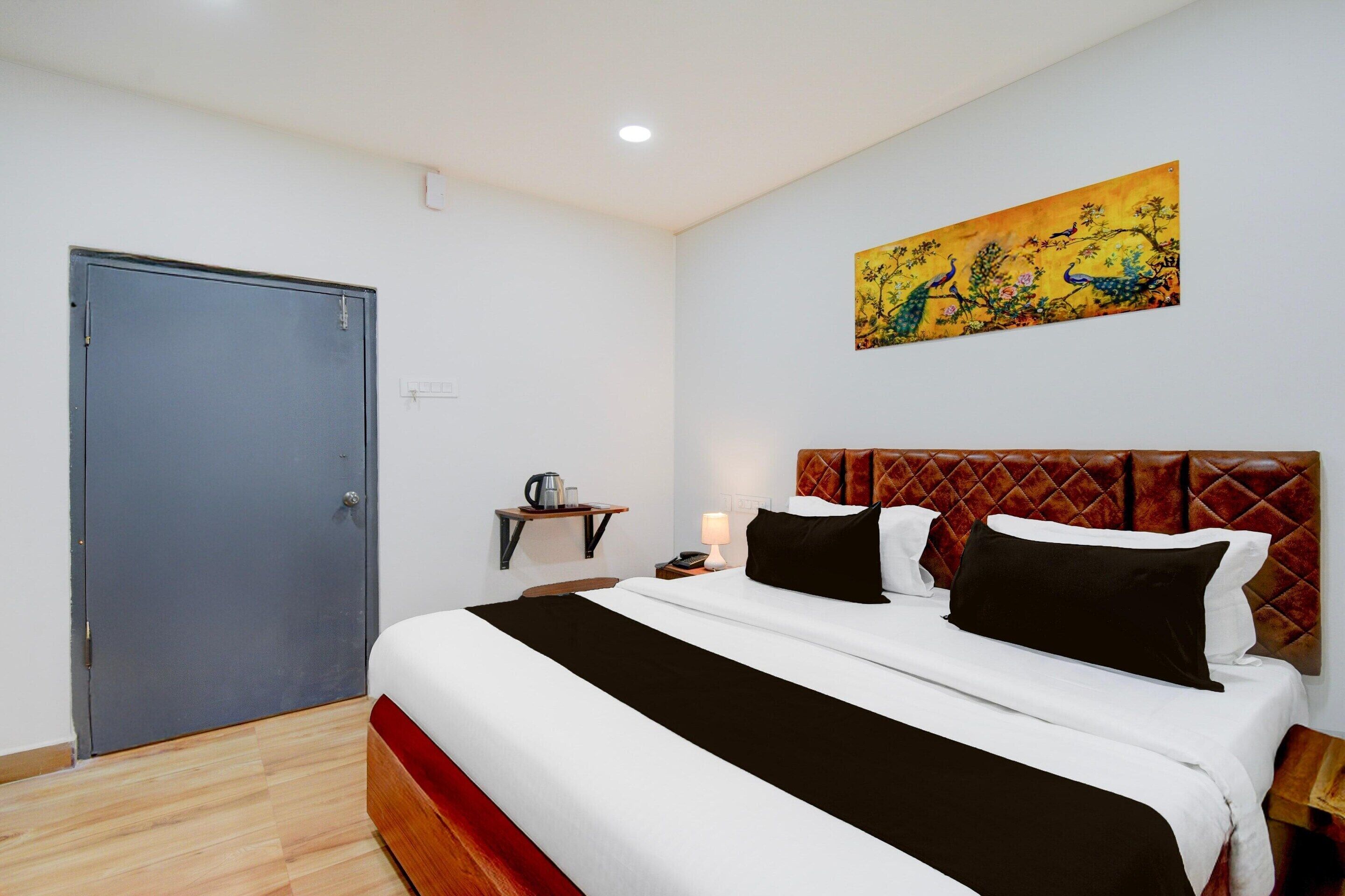 A hotel room with a bed, a door, a nightstand, a painting, and a wall socket.