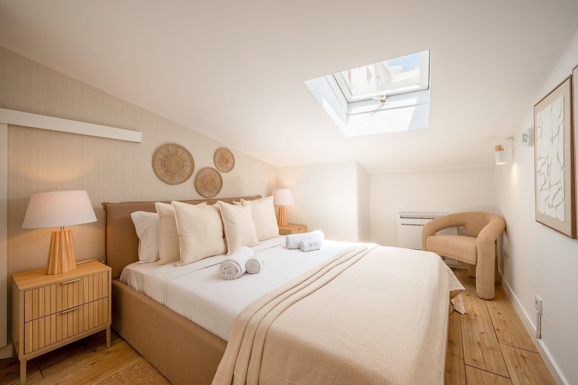 Un dormitorio con tragaluz, cama con ropa blanca, mesita de noche de madera, lámpara y aire acondicionado de pared.