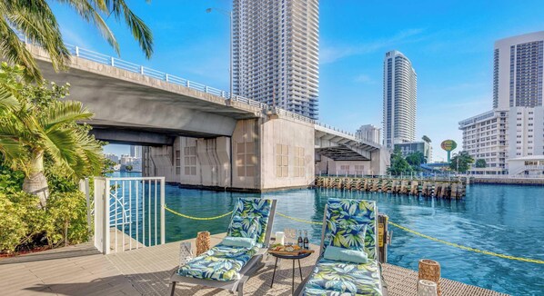 Zona de piscina frente al agua, con sillones, una mesa con bebidas y vista al horizonte de la ciudad.