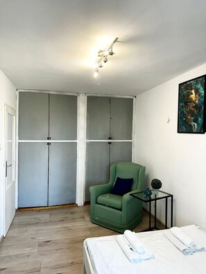 Ein Zimmer mit einem Bett, einem grünen Sessel, einem kleinen Tisch mit Pflanze und einem Kleiderschrank mit Leuchte darüber.