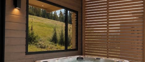 Un moderno espacio de spa con hidromasaje, escalones de madera y un gran ventanal con vistas a un paisaje verde.
