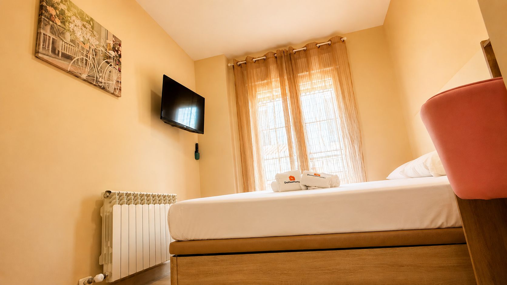 Ein Hotelzimmer mit einem Bett, einem an der Wand montierten Fernseher, einem Heizkörper und einem Fenster mit Vorhängen.