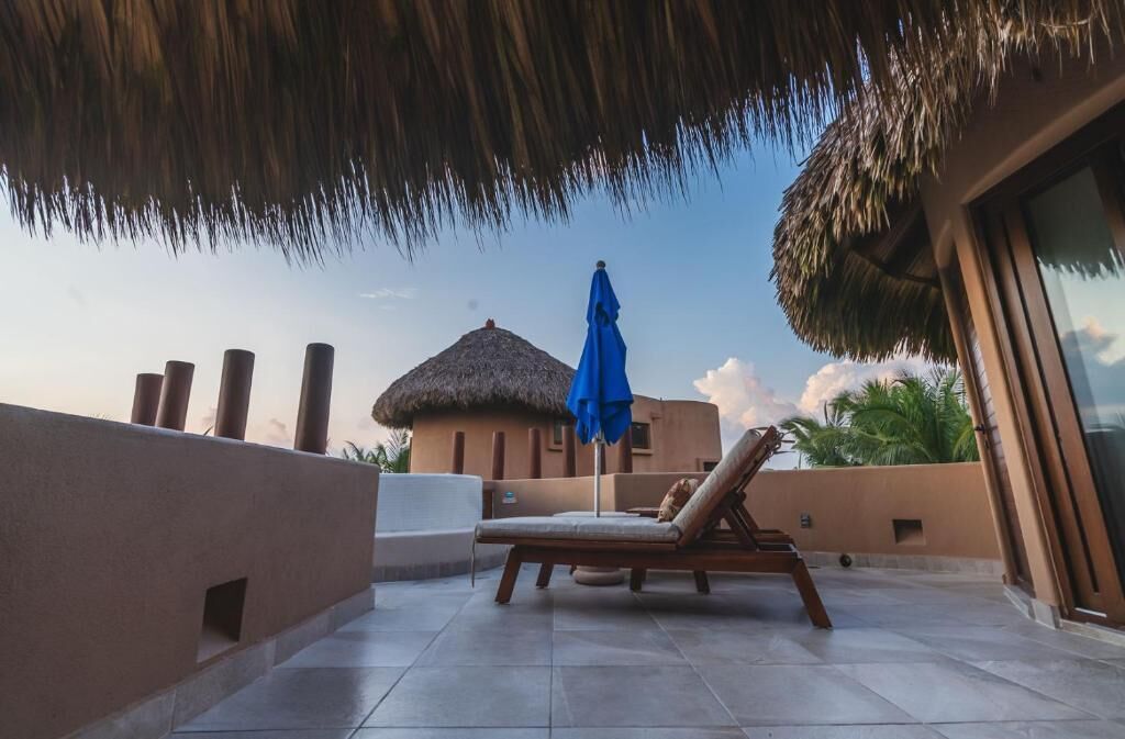 A rooftop terrace with a lounge chair and umbrella, a thatched-roof structure, and a view of palm trees.