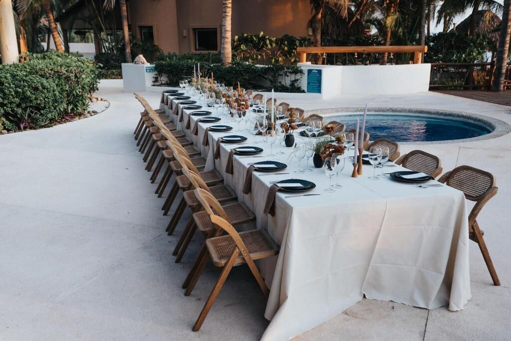 An outdoor dining area with a long table set for a meal, surrounded by chairs and a pool.