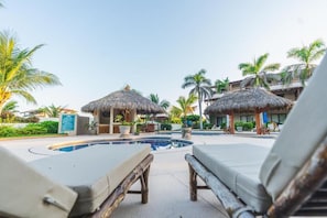 Zona junto a la piscina con sillones de descanso y estructuras de techo de paja, rodeada de palmeras y un cielo despejado.