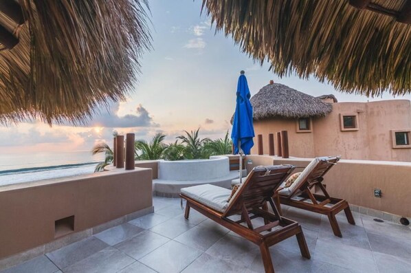 A terrace with lounge chairs, a thatched-roof building, and a view of the ocean.