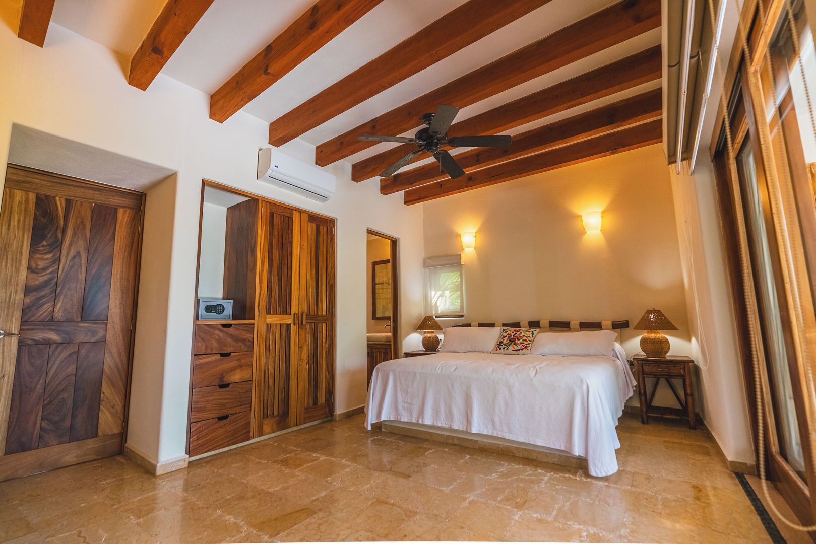 A bedroom with a bed, wooden ceiling beams, an air conditioning unit, a ceiling fan, and a nightstand with a lamp.