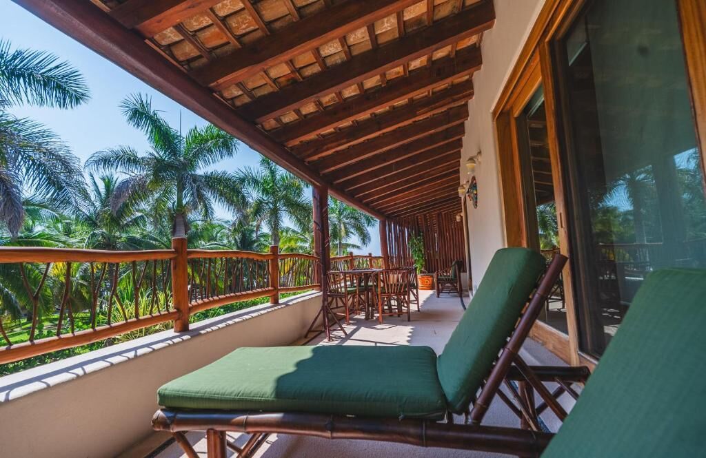 Un balcón con barandillas de madera, sillas con cojines verdes y vistas a palmeras.