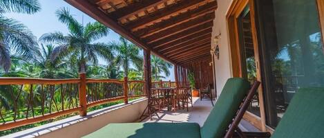 A balcony with wooden railings, green cushioned chairs, and a view of palm trees.