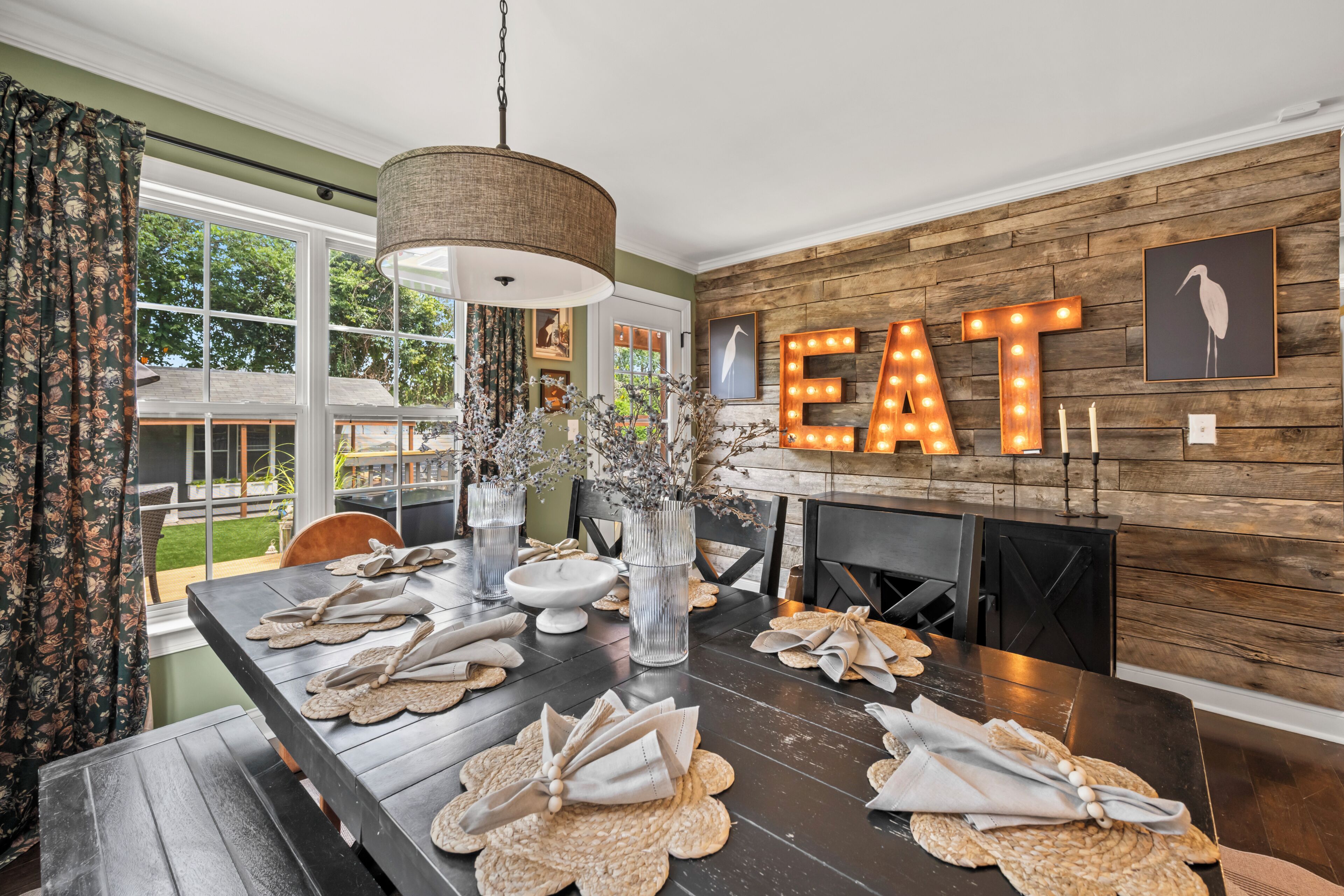 A dining area with a long table set for a meal, a large illuminated 'EAT' sign on the wall, and a view of the outdoors through large windows.