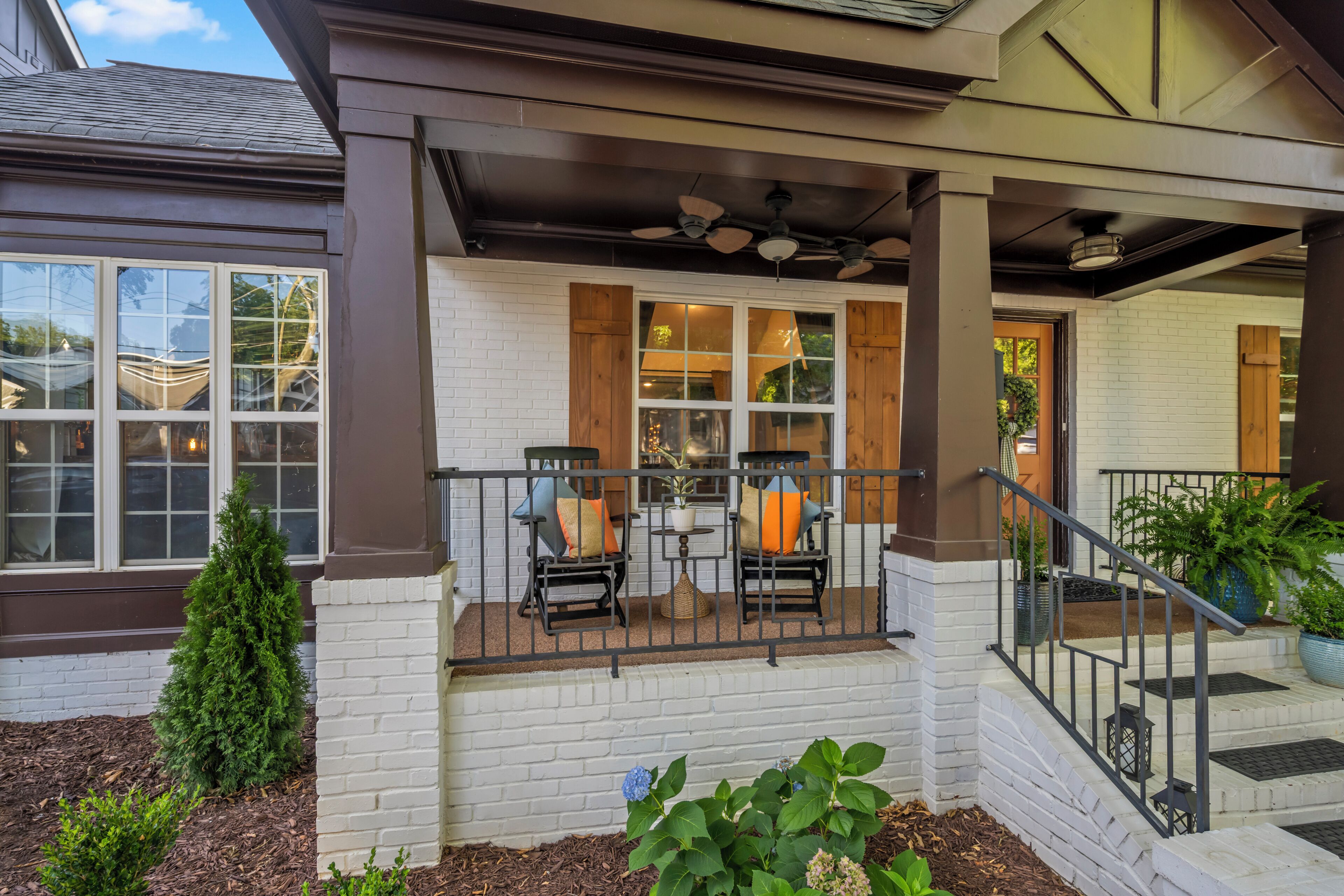 A covered porch with two chairs and a table, white brick walls, and a staircase leading to the entrance.