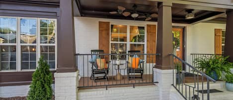 A covered porch with two chairs and a table, white brick walls, and a staircase leading to the entrance.