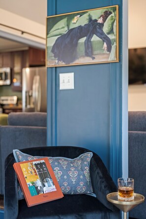 A room with a dark blue sofa, a framed picture on the wall, a small table with a glass of amber liquid, and a pillow with a floral pattern. - Escape to the Arts, Golf Sim, Theater, Hot Tub (Charlotte)