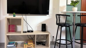 Olohuone, jossa seinälle on asennettu taulutelevisio, ja jossa on puinen hyllykkö, jossa on kirjoja ja elektronisia laitteita, pieni ruukkukasvi sekä ruokailutila, jossa on kaksi mustaa tuolia ja pöytä.