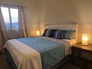 A neatly made bed with a headboard, two bedside lamps, and a window with curtains.