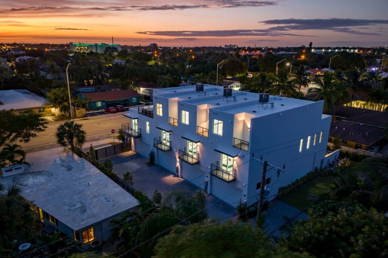 A modern multi-story building with balconies, surrounded by greenery and a view of a cityscape at dusk.