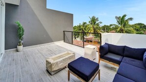 Ett modernt utomhusområde med sittmöbler, bestående av en soffa och ett fotstöd, en krukväxt och utsikt över palmer och byggnader.