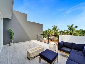 Ein moderner Außenbereich zum Sitzen mit einer Couch, einem Hocker, einer Topfpflanze und Blick auf Palmen sowie Gebäude.