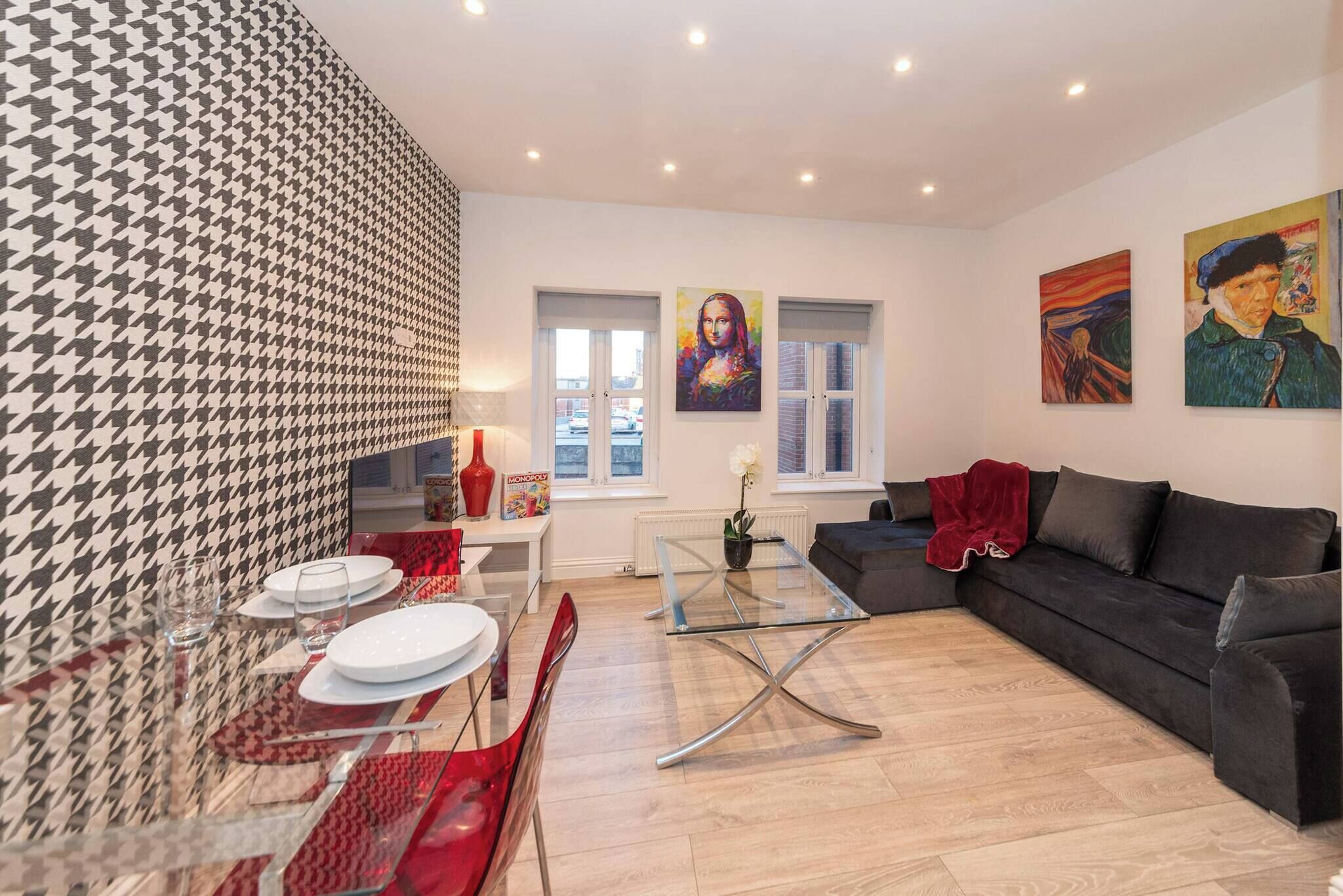 A modern living room with a glass coffee table, a black sofa, and a dining area with a red table runner.
