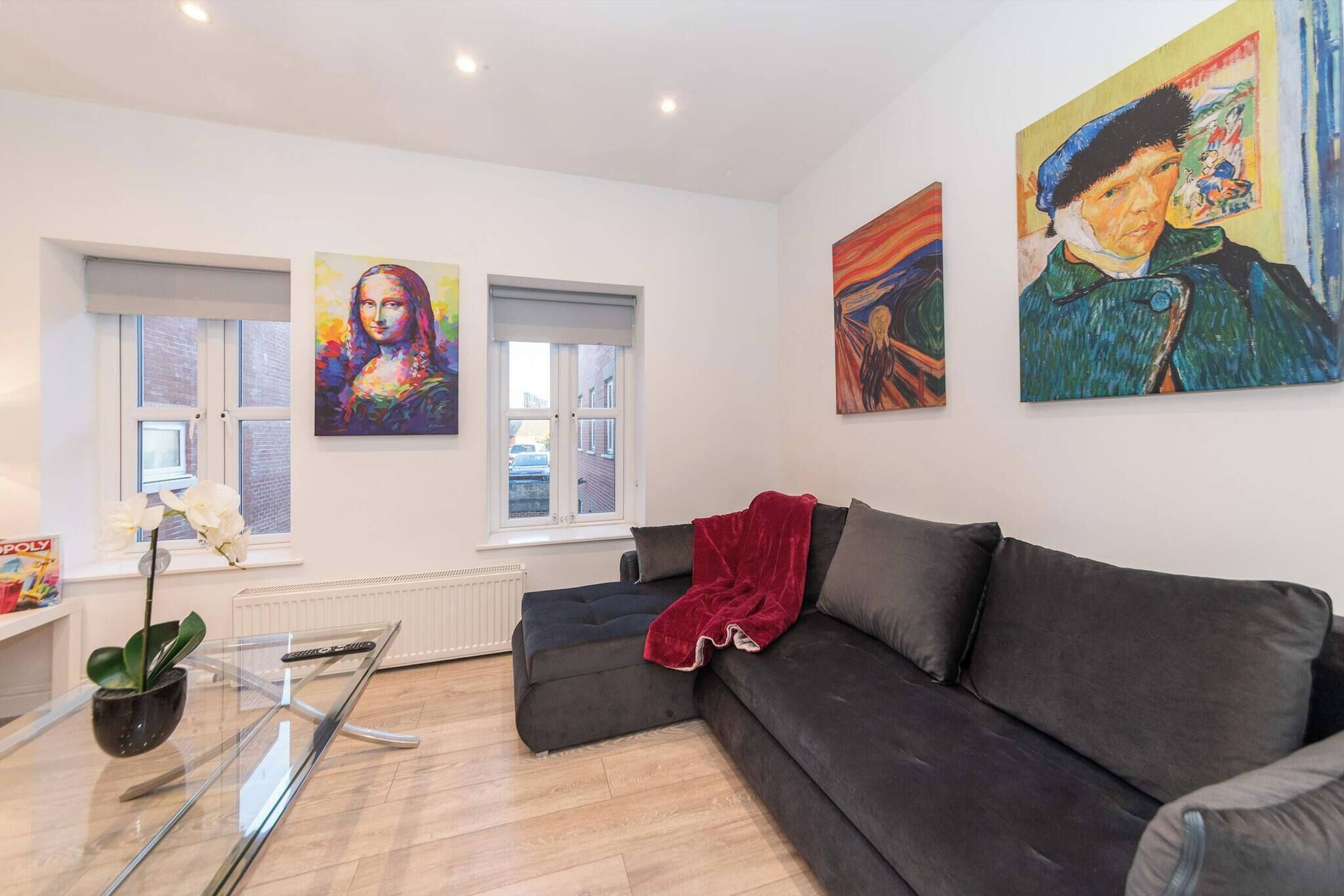A living room with a black sofa, a glass coffee table, a red throw blanket, and three paintings on the wall.