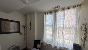 A bedroom with a bed, a standing fan, a heater, and a window with patterned curtains.